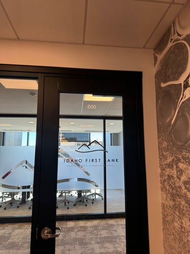 A glass office door with "IDAHO FIRST BANK" and its mountain logo etched in frosted glass. The number "600" is visible above the door. Inside, a conference room with chairs and a table can be seen.