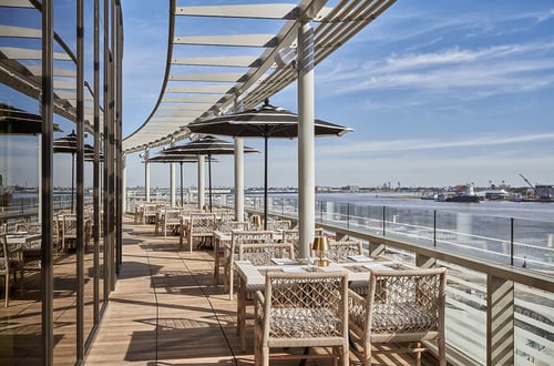 Outdoor restaurant terrace overlooking a calm river, featuring modern wicker chairs, tables with gold lamps, and large black umbrellas under a clear blue sky.