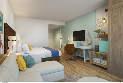 Hotel room with a chaise lounge, white bed facing a desk under a flat screen TV, and a white brick accent wall