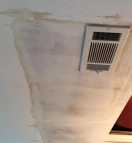 A textured ceiling showing significant water damage, with a large yellowish-brown stain spreading next to a rectangular HVAC ceiling vent.