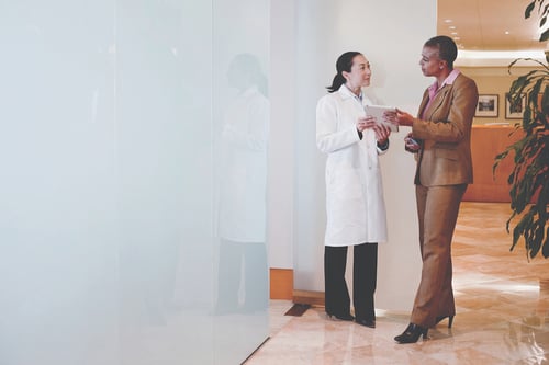 A doctor in a white coat and a businesswoman in a brown suit are engaged in a conversation in a bright, modern office. The mood is professional and collaborative.