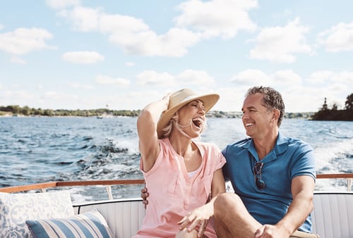 A cheerful couple enjoys a sunny day on a boat. The woman in a pink shirt and straw hat laughs, while the man in a blue shirt smiles back at her.