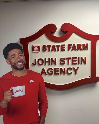 A person in a red sweater holds a mug labeled "JAKE" next to a State Farm sign for the John Stein Agency.