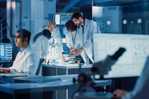Scientists in lab coats collaborate on equipment at workbench in modern blue-lit research laboratory.