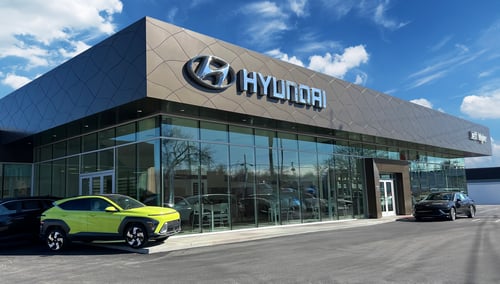 Glass building with a grey modern top. The Hyundai logo on the left and the Jeff Wyler logo on the right. A lime-green Hyundai-branded vehicle in the bottom-left corner. A black Hyundai vehicle in the bottom right corner.