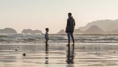 A man and child stand facing each other on a reflective beach at sunset, with gentle waves and distant hills creating a serene, warm atmosphere.
