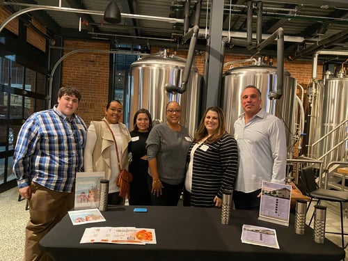 Six team members of A. E. Mazika Insurance Services standing together in a brewery with a table in front of them with flyers.