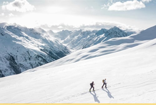 Two people hike up a snowy mountain under a bright sky with fluffy clouds. The expansive snow-covered peaks in the background convey a sense of adventure.