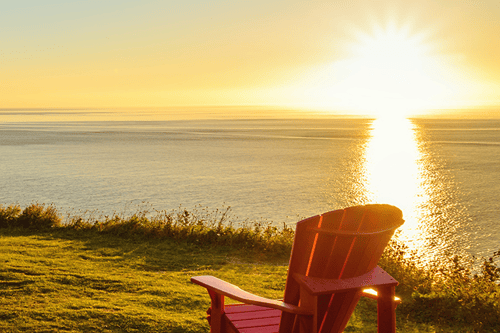 Chaise Adirondack rouge sur une falaise herbeuse surplombant la paisible baie côtière au coucher du soleil doré.