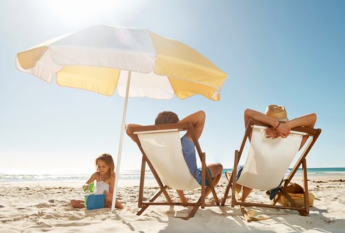 A child plays with a bucket and spade on a sunny beach, while two adults relax on deck chairs under a yellow umbrella, creating a peaceful scene.