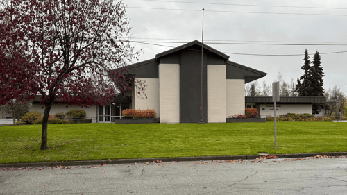 Front view of the Maplewood church building with tall light brick panels and dark trim. A red-leafed tree stands on the left. Both the north and south entrances are visible, with ramps and landscaped shrubs. A wide green lawn extends across the foregroun