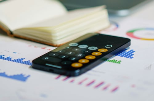 A smartphone with a calculator app open rests on financial charts, alongside an open notebook. The image conveys an analytical, professional tone.
