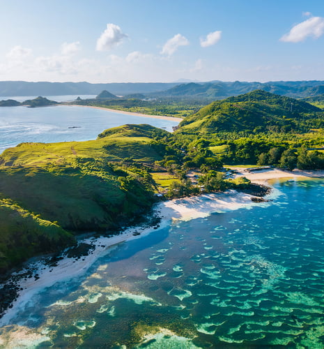 Aerial view of a lush, green coastal landscape with rolling hills, crystal-clear turquoise waters, and a pristine sandy beach under a bright blue sky.