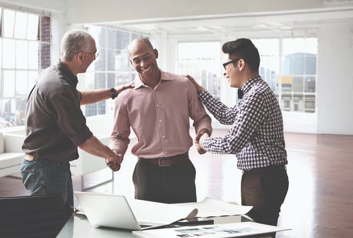 Three colleagues in a bright office celebrate with smiles and handshakes by a desk with a laptop and papers, conveying teamwork and success.
