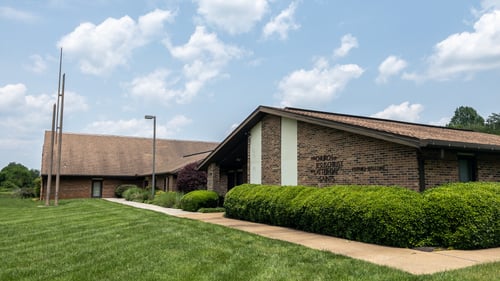 The Church of Jesus Christ of Latter-day Saints meetinghouse in Cleveland, Tennessee sits on a corner lot. The viewer is presented with a dark brick building and brown roofing, with the name of the Church mounted on the brick, beside white decorative paneling. A walkway leads to the front doors, wrapping around the landscaping comprised of manicured bushes. Three ornamental metal spires sit near the walkway,  in front of the building.