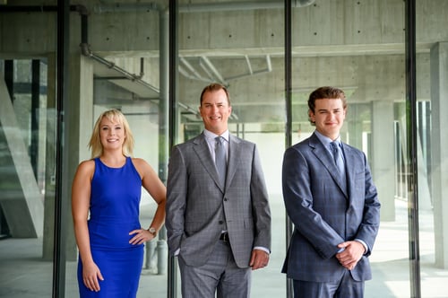 Emily left in blue dress, Kevin middle in grey suit, Carter right in blue suit standing in front of glass building