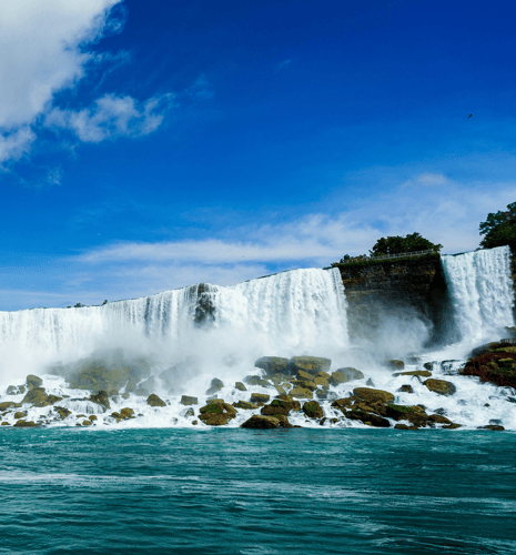A majestic waterfall cascades over a rocky cliff into a turquoise river below, surrounded by lush greenery. The sky is bright blue with scattered clouds.