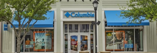 Photograph of Columbia Sportswear storefront with blue logo sign above the entrance. The large glass windows display mannequins dressed in outdoor clothing, and the store's interior is visible. The suite number 240 is shown on the door.