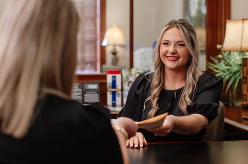 A smiling Bryant Bank employee with blonde hair, wearing a black top, hands a document to a customer over a dark wood desk. The office background is warm and professional with classic lamps and wooden trim.