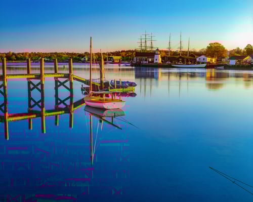 Calm harbor at sunset with sailboats moored at wooden dock, reflecting autumn foliage and historic tall ships in still water, featuring lighthouse and waterfront buildings along shoreline.