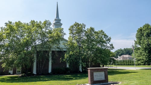 The Church of Jesus Christ of Latter-day Saints meetinghouse in Ooltewah, Tennessee. The viewer is presented with a red brick building, pitched roof and one white spire. Four large white columns decorate the front of the building, with two trees planted directly in front of them. A red brick marker with the name of the Church engraved in granite sits a few yards in front of the white-columned building front.
