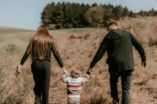 A family of three walks hand-in-hand through a grassy field, with a clear blue sky and trees in the background.