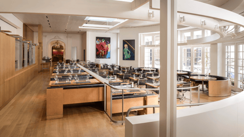 A modern restaurant dining area with sleek wooden tables and glassware set for service. Large windows allow natural light, and vibrant artwork decorates the walls.