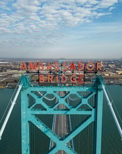 Aerial view of the Ambassador Bridge with turquoise steel framework spanning a river, with red letters spelling "AMBASSADOR BRIDGE" on the tower, urban landscape and cloudy blue sky in background