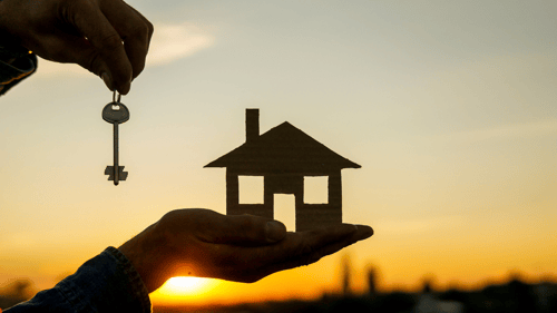 person holding a key and small model home during sunset