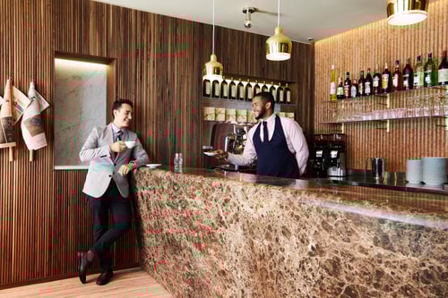 Man holding a cup of coffee speaking with a barista at a cafe counter.