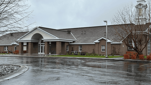 Church building with brown brick walls, peaked rooflines, and a prominent steeple, seen from a wet parking lot.