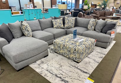 Contemporary gray sectional sofa with oversized patterned ottoman and accent pillows at Slumberland Furniture Burlington. Discover comfortable sectionals and living room furniture in Burlington, Iowa.
