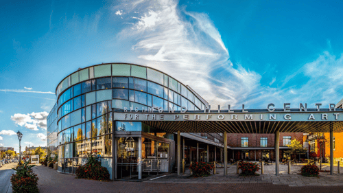 Modern performing arts center with a curved glass facade under a vibrant blue sky and swirling white clouds. The architecture feels open and inviting.