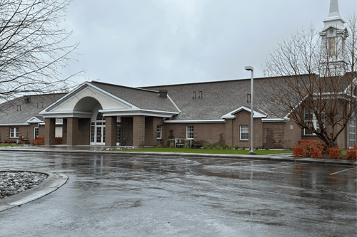 Church building with brown brick walls, peaked rooflines, and a prominent steeple, seen from a wet parking lot.