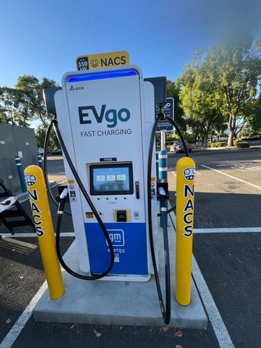 NACS EVgo fast charging unit installed in marked parking space at a public charging station, with EVgo logos on the charger,  and yellow protective bollards around the unit.