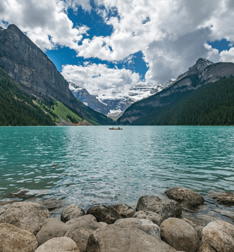 Two people ride a canoe in the distance, along a rocky shoreline with mountains in the background.