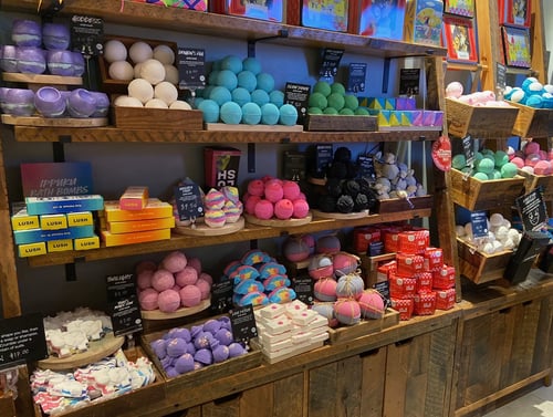 Large display of wall mounted unpackaged brightly coloured bath products in wooden pens creating mounds of colour.