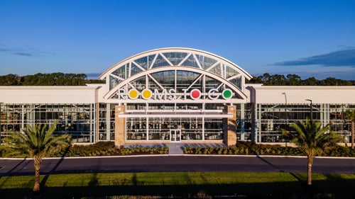 Aerial view of the Rooms To Go store entrance with a large, curved glass facade and prominent "Rooms To Go" signage. The building is surrounded by palm trees and landscaped greenery.