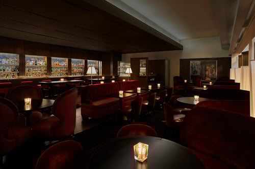 A dimly lit lounge bar with rich red velvet seating, dark wooden tables, and candlelit ambiance. The back wall displays a wide variety of bottles, creating a cozy, intimate setting.