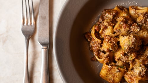 Plate of rigatoni pasta with meat sauce and grated cheese next to a fork and knife on a marble surface