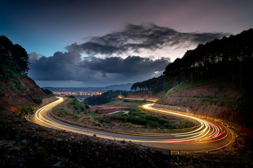 A long-exposure night photograph of a winding mountain road with glowing vehicle light trails curving through a valley, forested hillsides, and a dramatic stormy sky above a distant city.