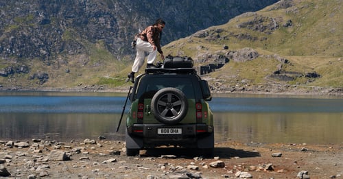 Rear view of a green Defender parked by a mountain lake with a person loading gear on the roof.