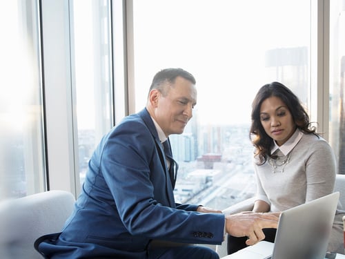 Man in a blue suit and woman in a gray sweater collaborate on a laptop in a modern office with large windows overlooking a cityscape.
