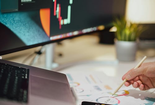 A close-up of a person working at a desk. A computer screen displays a stock chart. Nearby, a hand holds a stylus over printed graphs, and a small plant is visible, creating a focused and analytical atmosphere.