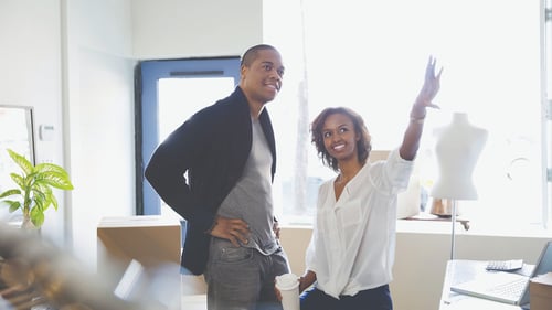 Two people in a bright space discuss ideas. A woman, smiling, gestures upward with enthusiasm, holding a coffee cup. A man stands beside her, listening intently.