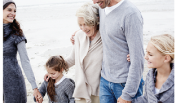 A multigenerational family enjoys a walk on a beach. They are smiling, holding hands, wearing cozy sweaters, and surrounded by a peaceful, overcast sky.