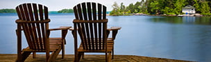 Two wooden chairs on a lakeside deck overlook calm water, surrounded by lush trees and a distant white house, conveying tranquility and relaxation.