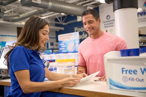 A Leslie’s employee in a blue shirt helping a customer at the counter with pool maintenance products visible in the background.