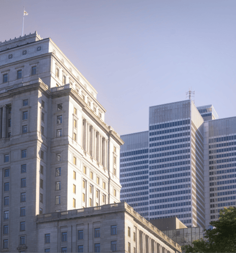 Un bâtiment classique en pierre, à l'architecture détaillée, est baigné par une douce lumière. À l'arrière-plan, des gratte-ciel modernes en verre se dressent sous un ciel dégagé, créant un contraste saisissant entre l'ancien et le nouveau.