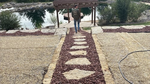 Owner Alex of Simply Snow & Lawn walking a stone stepping path with red lava rock toward a wooden pergola and pond in McFarland, WI